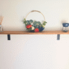 A solid oak floating shelf with black metal L-shaped brackets holding a decorative floral wreath, a reed diffuser, and a blue wax melt warmer against a light wall.