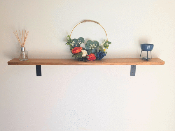 A solid oak floating shelf with black metal L-shaped brackets holding a decorative floral wreath, a reed diffuser, and a blue wax melt warmer against a light wall.
