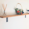 Warm-toned solid oak shelf with a softened front edge, supported by minimalist black industrial brackets, displaying home décor items like a scent diffuser and a decorative ring with faux flowers